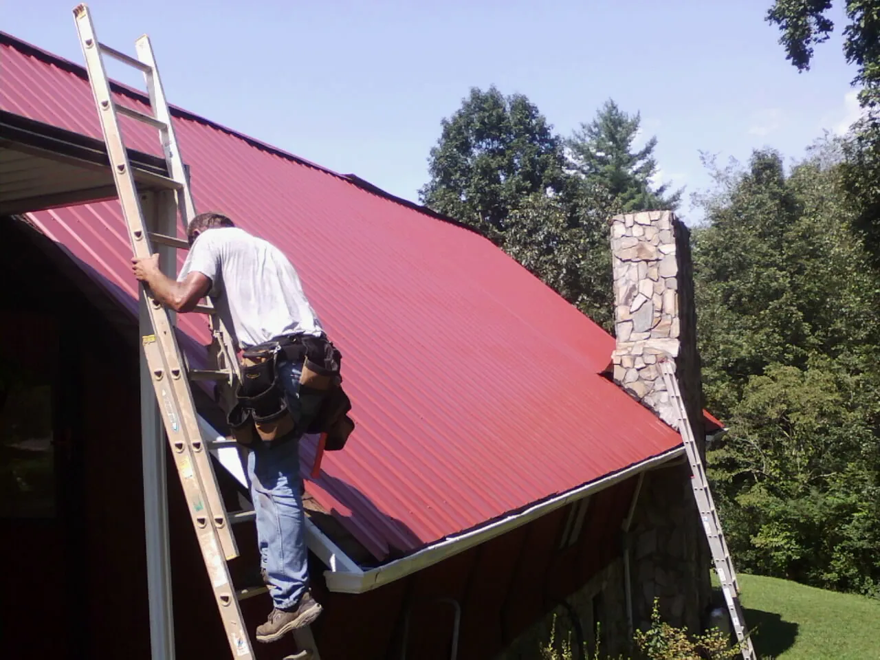 Roofer on a ladder beside a red metal roof and stone chimney — Soffit & Fascia Repair by Roofing Brothers in Center Tuftonboro