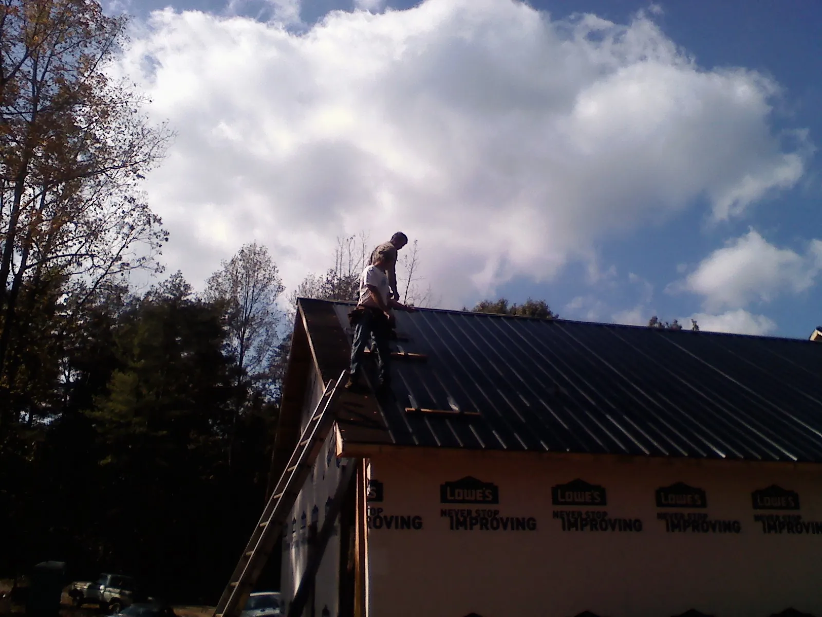 Roofer fastening dark metal panels high against a cloudy sky, from a Roof Inspection job in Center Tuftonboro